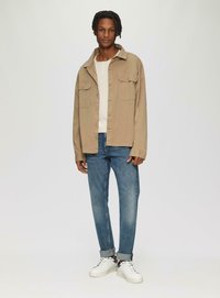 Beige Hemd mit Knöpfen und zwei Brusttaschen, über einem cremefarbenen Oberteil getragen, kombiniert mit blauen Jeans und weißen Sneakers mit schwarzen Akzenten.