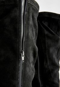Bottes noires en daim montantes avec une fermeture éclair sur toute la longueur, des accessoires en métal argenté brillant, et une texture douce avec des détails de surpiqûres contrastants.