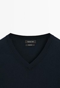 Baumwoll-Pullover mit V-Ausschnitt in dunklem Marineblau, mit gerippten Details am Kragen und einem Etikett, das die Marke und den Materialinhalt angibt.