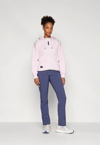 Felpa con cappuccio rosa chiaro con zip e coulisse, dettagli neri, abbinata a pantaloni blu navy. Il modello indossa scarpe da ginnastica bianche con dettagli colorati.