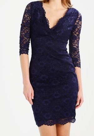 Femme portant une robe en dentelle bleu marine moulante, avec des manches trois-quarts et un décolleté en V.