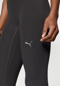 Zbliżenie osoby noszącej czarne leginsy Puma z widocznym logo Puma w pobliżu uda i dłonią osoby spoczywającą obok nich.