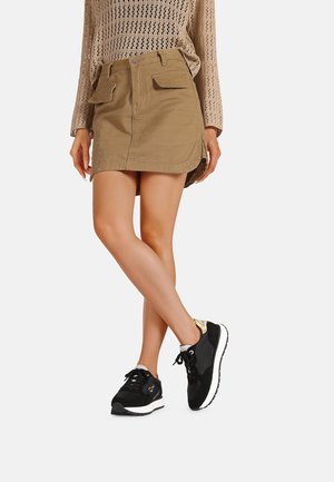 Gonna mini khaki con tasche frontali, abbinata a sneakers nere con un accento riflettente, su uno sfondo bianco semplice.
