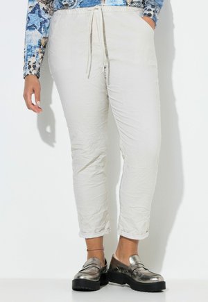 Person iført krøllede, off-white bukser med snor, metallic loafers og en blå mønstret bluse med lange ærmer, stående op ad en hvid væg.