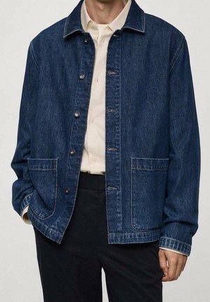 Mann trägt dunkelblaue Jeansjacke über cremefarbenem Hemd mit Knöpfen und schwarzen Hosen, Hände locker an den Seiten.