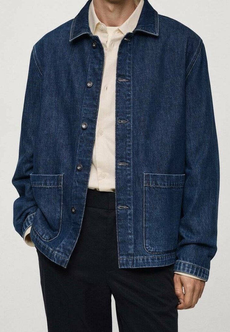 Homme portant une veste en jean bleu foncé sur une chemise crème à boutons et un pantalon noir, mains détendues sur les côtés.