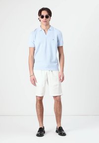 Polo din tricot de culoare albastru deschis cu mâneci scurte, pantaloni scurți din in alba, pantofi slip-on negri și ochelari de soare rotunzi negri. Un ansamblu simplu și casual.