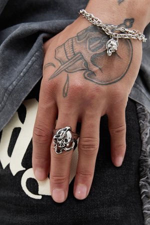 Bague en argent avec un design détaillé de crâne entrelacé de serpents, portée sur une main avec un tatouage visible, sur fond de tissu sombre.