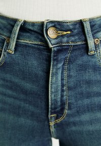 Niebieskie dżinsy z mosiężnym guzikiem z logo "Lee", kontrastującym żółtym przeszyciem i klasycznym pięciopocketowym designem.