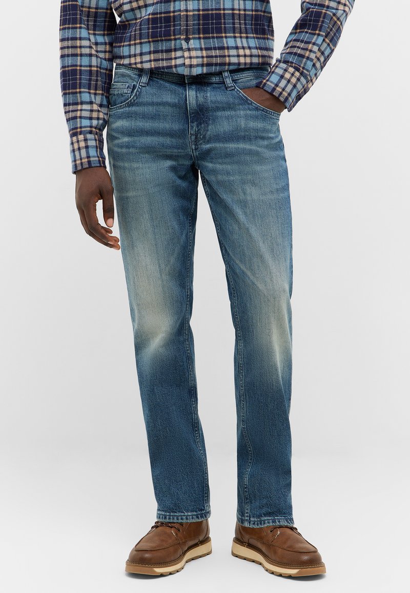 Męskie niebieskie dżinsy z przetarciami, pięcioma kieszeniami, prostym krojem nogawki i subtelnym efektem przecierania na przodzie. Nosi się je z brązowymi butami.