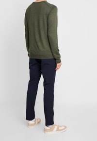 Grüner Langarmpullover mit Rundhalsausschnitt, kombiniert mit navyblauen Hosen und beigen Sneakers, die weiße Akzentdetails und einen strukturierten Stoff aufweisen.