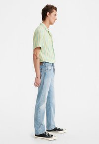 Joven de pie de lado, vistiendo una camisa de rayas amarilla y azul de manga corta, jeans de color azul claro y zapatillas negras.