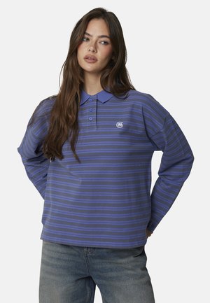 Giovane donna con lunghi capelli castani che indossa una maglietta polo a maniche lunghe a righe blu e nere e jeans blu, in piedi con le mani sui fianchi.