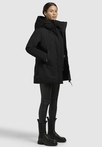 Sort pufferjakke med stor krave, sidelommer og lynlås. Sammen med sorte leggins og ankelstøvler, på en neutral baggrund.