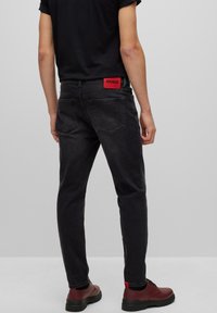 Černé džínové kalhoty slim fit, s červenou nášivkou "HUGO" na pase a dvěma zadními kapsami. Nosené s vínovými botami.
