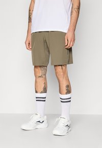 Bermudas caqui feitas de um tecido leve apresentam bolsos laterais, combinadas com ténis brancos e meias brancas às riscas.