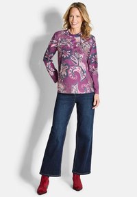 Frau lächelt, trägt ein lila florales Langarmoberteil, dunkelblaue Schlagjeans und rote Stiefel und steht vor einem weißen Hintergrund.