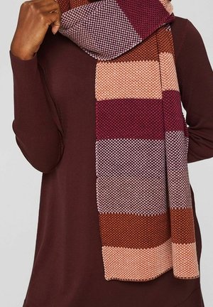 Personne portant une écharpe en tricot à larges rayures horizontales bordeaux, rose, lavande et marron, par-dessus un haut à manches longues couleur marron foncé.