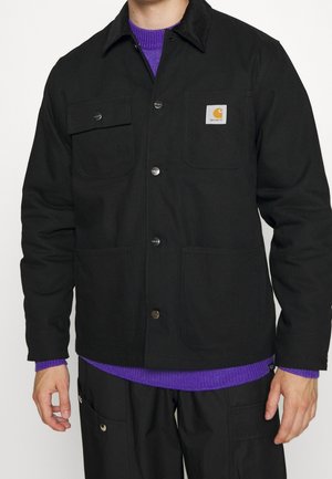 Person trägt eine schwarze Carhartt-Jacke über einem lila Strickpullover, dazu schwarze Hosen mit geknöpften Taschen.