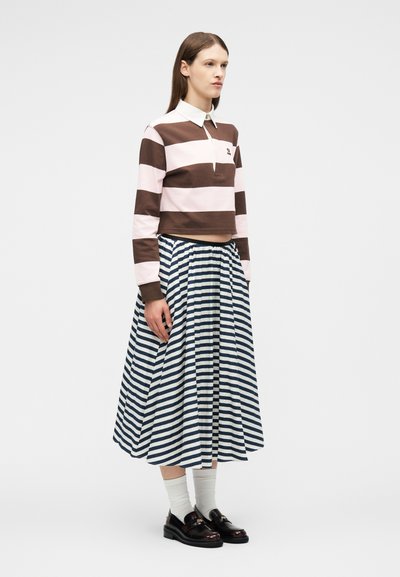 Γυναίκα που φοράει μακρυμάνικη μπλούζα πόλο με καφέ και ανοιχτό ροζ ρίγες, μίντι φούστα με οριζόντιες ρίγες σε μπλε ναυτικό και λευκό χρώμα, άσπρες κάλτσες και μαύρα μοκασίνια, όρθια.