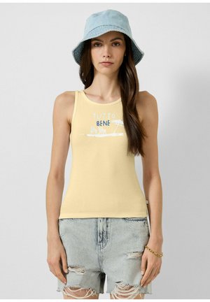 Femme portant un haut sans manches jaune avec le texte « Tutto Bene », un chapeau bob bleu clair et un short en jean clair déchiré, debout devant un fond uni.