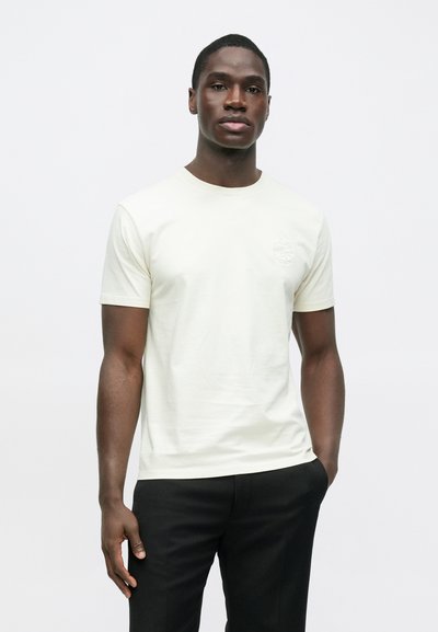 Camiseta de manga corta color crema claro con cuello redondo, con un logo sutil en el pecho izquierdo. Se lleva con pantalones negros, textura lisa.