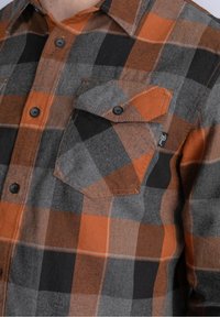 Koszula flanelowa w kratę w kolorach szarym, pomarańczowym i czarnym. Posiada zapinaną na guziki kieszeń na piersi oraz czarne guziki. Miękka w dotyku o luźnym kroju.