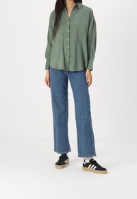Chemise verte à boutons, avec une coupe décontractée, assortie à un jeans bleu taille haute. Des baskets noires avec des rayures blanches et des semelles en caoutchouc complètent le look.