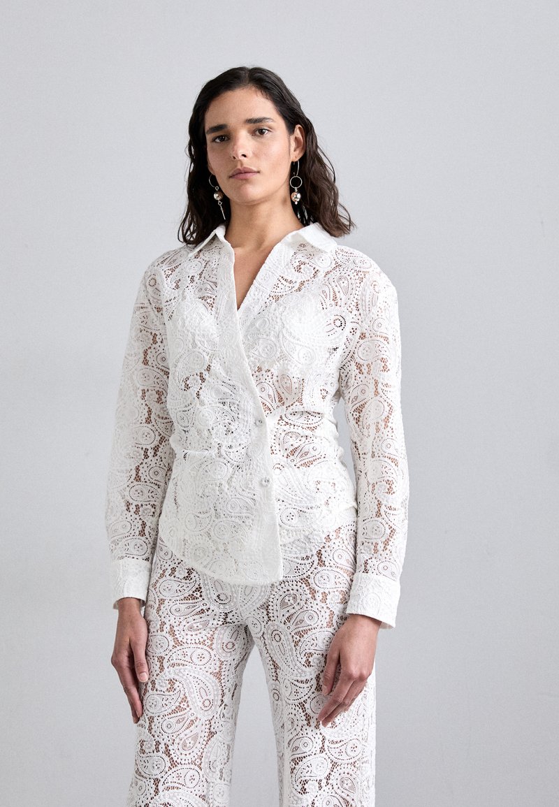 Chemise blanche en dentelle à boutonnière avec des motifs floraux complexes, associée à un pantalon en dentelle assorti. Dispose d'un col et de manches longues.
