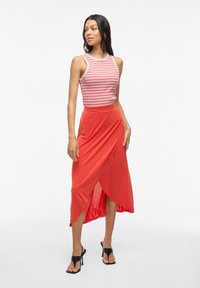 Top sin mangas a rayas con líneas rojas y blancas, combinado con una falda coral fluida. La falda tiene un diseño envolvente y una abertura alta. Sandalias de tacón negras.