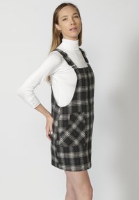 Čiernobiele kockované overall šaty s priliehavým strihom, nastaviteľnými ramienkami a bočnými vreckami. Nosené cez biely rolák.