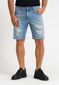 Shorts de mezclilla azul claro con detalles desgastados, que presentan desgastes y bordes deshilachados, terminados con un clásico diseño de cinco bolsillos. Zapatillas negras completan el look.