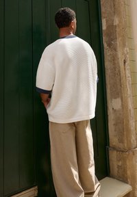 White textured shirt with navy accents and wide sleeves, paired with beige loose-fitting pants. Standing against a green door.
