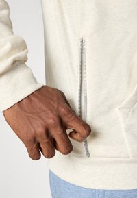 Ruka drží strieborný zips na svetlo béžovom mikine, ktorá je kombinovaná so svetlo modrými nohavicami.