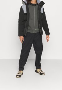 Chaqueta tipo plumón con paneles en gris y negro, cierre con cremallera, capucha ajustable; combinada con pantalones oscuros y zapatillas beige y negras.