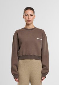 Felpa corta marrone in materiale morbido con maniche lunghe e polsini a costine. Presenta un piccolo logo bianco sul petto a sinistra.