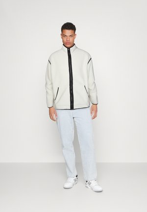 Fleecejakke i hvid med sort lynlås foran, med sidelommer og sorte kanter. Sammen med lyseblå jeans og hvide sneakers.
