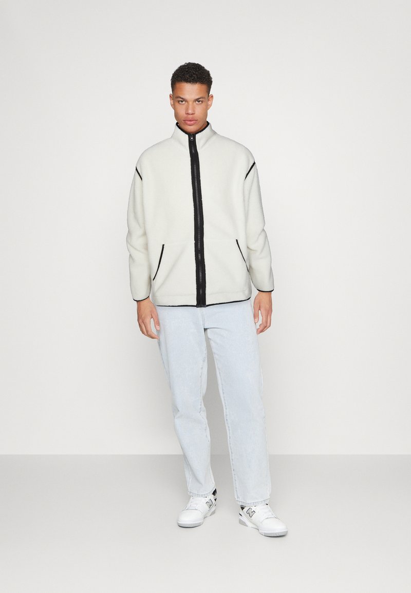 Fleece jack in het wit met een zwarte rits aan de voorkant, met zijzakken en zwarte randen. Gecombineerd met lichtblauwe spijkerbroeken en witte sneakers.