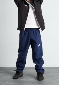 Pantalones deportivos azul marino con cintura elástica y diseño de tres rayas, combinados con una chaqueta negra con detalles en blanco y una camiseta blanca debajo.