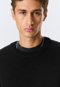 Jeune homme aux cheveux courts et bouclés portant un pull en tricot noir par-dessus une chemise noire, faisant face à l'avant avec une expression neutre.