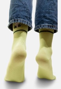 Jasnożółte skarpetki w prążki z zielonym uśmiechniętym motywem, noszone z podwiniętymi niebieskimi dżinsami, pokazując miękką teksturę i kształt.