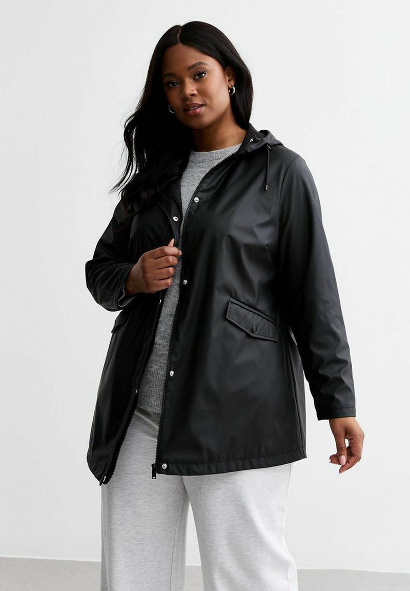 Svart vattentålig jacka med dragsko-hood, tryckknappar, framficka och avslappnad passform; bärs över en grå tröja och ljusa byxor.