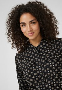 Schwarzes Hemd mit Knopfleiste und rundem Muster in Weiß und Gold, mit zwei Brusttaschen und Kragen. Der Stoff wirkt glatt und leicht.