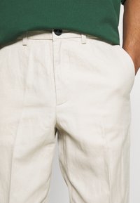 Pantalon beige en lin léger avec poches avant, fermeture à boutons, et texture lisse, associé à un t-shirt vert.