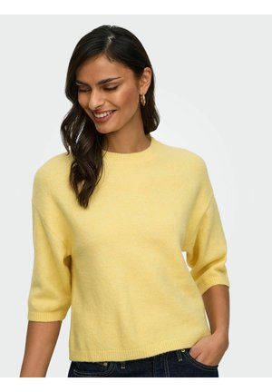 Femme aux cheveux foncés portant des boucles d'oreilles créoles en or et un pull jaune à manches courtes, souriante et regardant vers le bas, main dans la poche du jean.