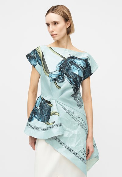 Γυναίκα που φοράει ασύμμετρο ανοιχτό μπλε τοπ με μεγάλα σκούρα λουλουδένια σχέδια και τυπωμένο κείμενο, συνδυασμένο με λευκή φούστα.