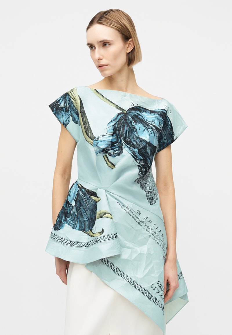 Frau trägt asymmetrisches hellblaues Oberteil mit großen dunklen Blumenmustern und gedrucktem Text, kombiniert mit einem weißen Rock.