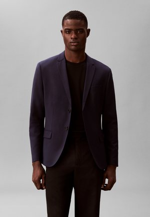 Homme debout face à l’avant portant un blazer bleu marine foncé sur une chemise noire et un pantalon noir sur un fond gris uni.