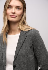 Szary, teksturowany blezer o dopasowanym kroju, z klapą z wycięciem i pojedynczym zapięciem na guzik, noszony na białej bluzce.