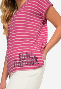 Camiseta a rayas rosa y gris con cuello en V y mangas enrolladas, que presenta el texto "fuld nordisk" en negro. Llevada sobre pantalones blancos.